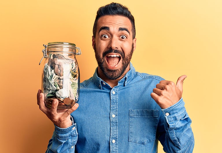 Delighted man holding a huge jar of cash in one hand while giving the thumbs up with the other