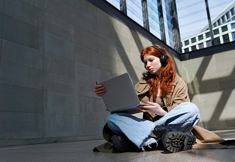 A young woman wearing headphones is sitting cross-legged on the floor looking at a laptop